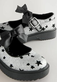 Des chaussures Mary Jane en paillettes argentées avec des motifs d'étoiles noirs, des nœuds en satin noir sur la sangle et une semelle en caoutchouc noir robuste.