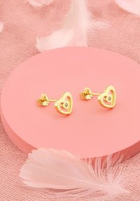 Gul hjärtformade studörhängen med en central sten, placerade mot en rosa cirkulär displayyta och omgivna av mjuka fjädrar.