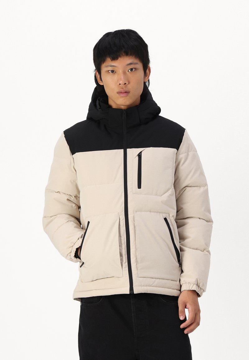 Beige und schwarze Pufferjacke mit Kapuze, ausgestattet mit einem Frontreißverschluss, zwei Seitentaschen und einer Brusttasche mit Reißverschluss.
