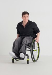 Mann med kort brunt hår som har på seg en svart skjorte og grå bukser, skyver en sort rullestol med grønne detaljer foran en ensfarget hvit bakgrunn.