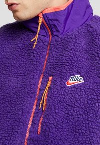 Pullover in pile viola con zip arancione e cordini, caratterizzato da un logo Nike sul petto. Materiale testurizzato con colletto alto.