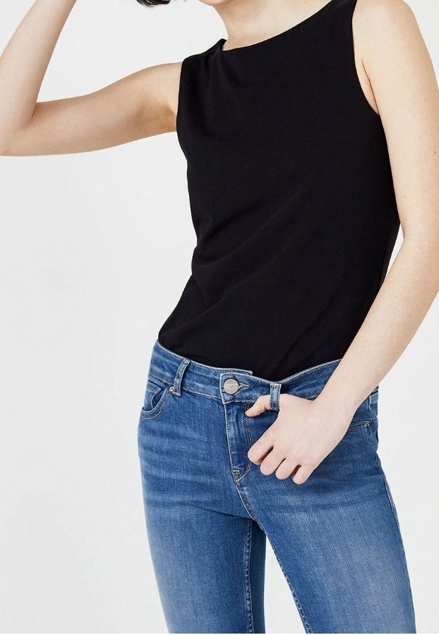 Kvinna som bär en ärmlös svart topp och blå jeans, med en hand som justerar jeansens midja.