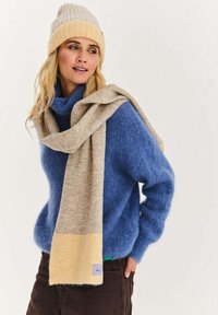 Niebieski, puchaty sweter, szalik w blokowe pasy w kolorach beżowym i żółtym zarzucony na ramiona, jasnobeżowa dziergana czapka, brązowe spodnie, swobodny strój.