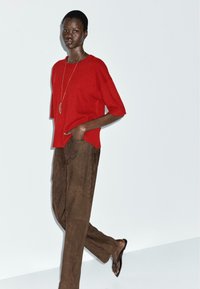 Pullover rosso a maniche corte lavorato a maglia abbinato a pantaloni larghi in suede marrone. Presenta una sottile rigatura verticale e una vestibilità rilassata.