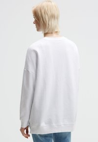 Frau mit kurzem blonden Haar, die einen schlichten weißen übergroßen Pullover und eine blue Jeans trägt, nach links blickend vor einem hellgrauen Hintergrund.