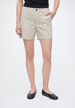 Vrouw draagt beige korte broek met zwarte verticale spijkerstrepen, zwarte platte schoenen en een zwart top met één hand in de zak.