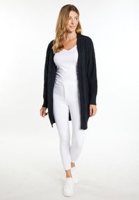 Zwarte gebreide cardigan met geribbelde textuur en kabelbreipatronen, gedragen over een witte top en witte slim-fit broek, gecombineerd met witte sneakers.