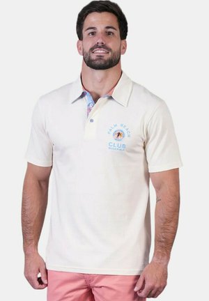Homme portant un polo crème avec le logo "Palm Beach Club Buckfield" et un short rose, debout devant un fond uni.
