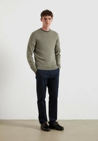 Męski sweter w oliwkowym kolorze z prążkowanym dekoltem, w komplecie z ciemnonawygodnymi spodniami i czarnymi butami. Prosty design, miękka tekstura. Bez wzorów ani akcentów.