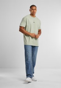 Light green cotton t-shirt with short sleeves, features a central black printed line, worn with light blue denim jeans and white sneakers.