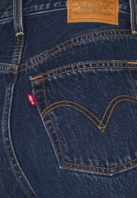 Vaqueros de mezclilla oscura con un patrón de costuras en cobre prominente, un parche de cuero con la marca y una etiqueta roja de Levi's en el bolsillo trasero.