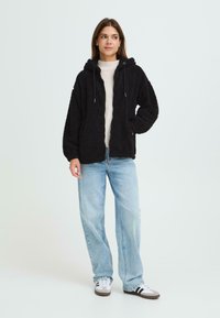 Hoodie en polaire noire et fluffy avec cordons de serrage, poches avant et coupe décontractée, associé à un jean bleu clair et des baskets.