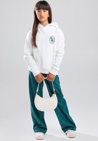 Weißer Hoodie mit einer Fronttasche und grafischem Druck, kombiniert mit türkisfarbenen, weit geschnittenen Hosen und einer kleinen weißen Handtasche. Weiße Sneakers runden das Outfit ab.