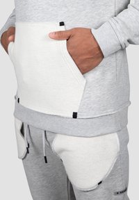 Sudadera gris con un diseño de bolsillo texturizado en blanco y acentos en negro. La tela es suave, con puños de canalé y un corte relajado.