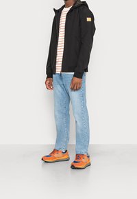Giacca nera con zip e cappuccio, maglietta a righe bianche e rosse, jeans in denim azzurro chiaro e scarpe da ginnastica arancioni.