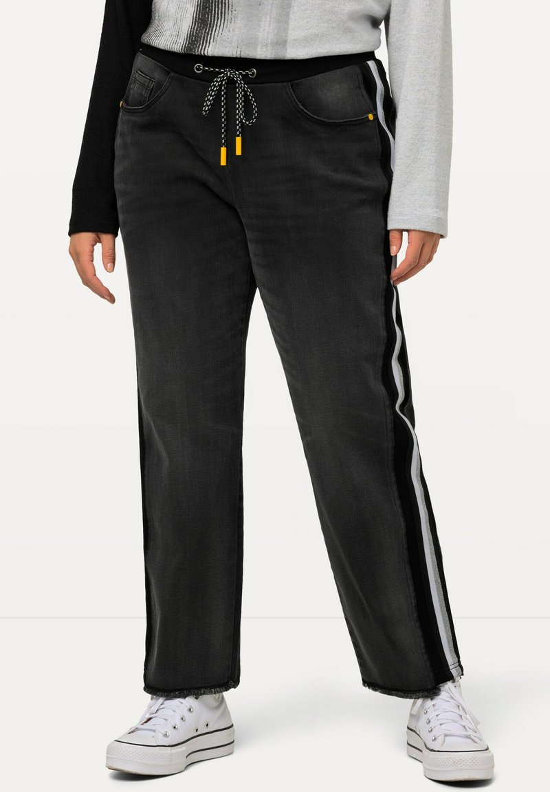 Pantalones de mezclilla negros con cintura ajustable, rayas laterales en blanco y negro, y dobladillos deshilachados. Combinados con zapatillas blancas.