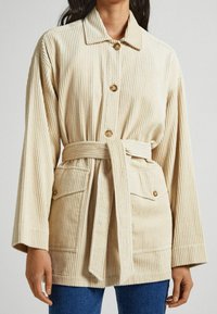 Giacca in corduroy beige con colletto, bottoni frontali, cintura a fusciacca e tasche applicate. Il tessuto ha una texture rigata e presenta maniche larghe.