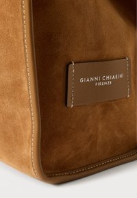 Rjava nubuk torbica z gladkimi usnjenimi poudarki, šivi na robovih in označba z napisom "Gianni Chiarini" v beli barvi.