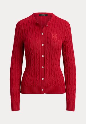 Femme portant un cardigan rouge à torsades, un pantalon blanc à jambes larges, et tenant un petit sac rouge avec une bandoulière en chaîne.