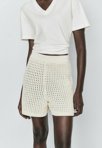 Cream-colored knitted shorts with a textured, open-weave pattern and elastic waistband, paired with a white cropped V-neck t-shirt.