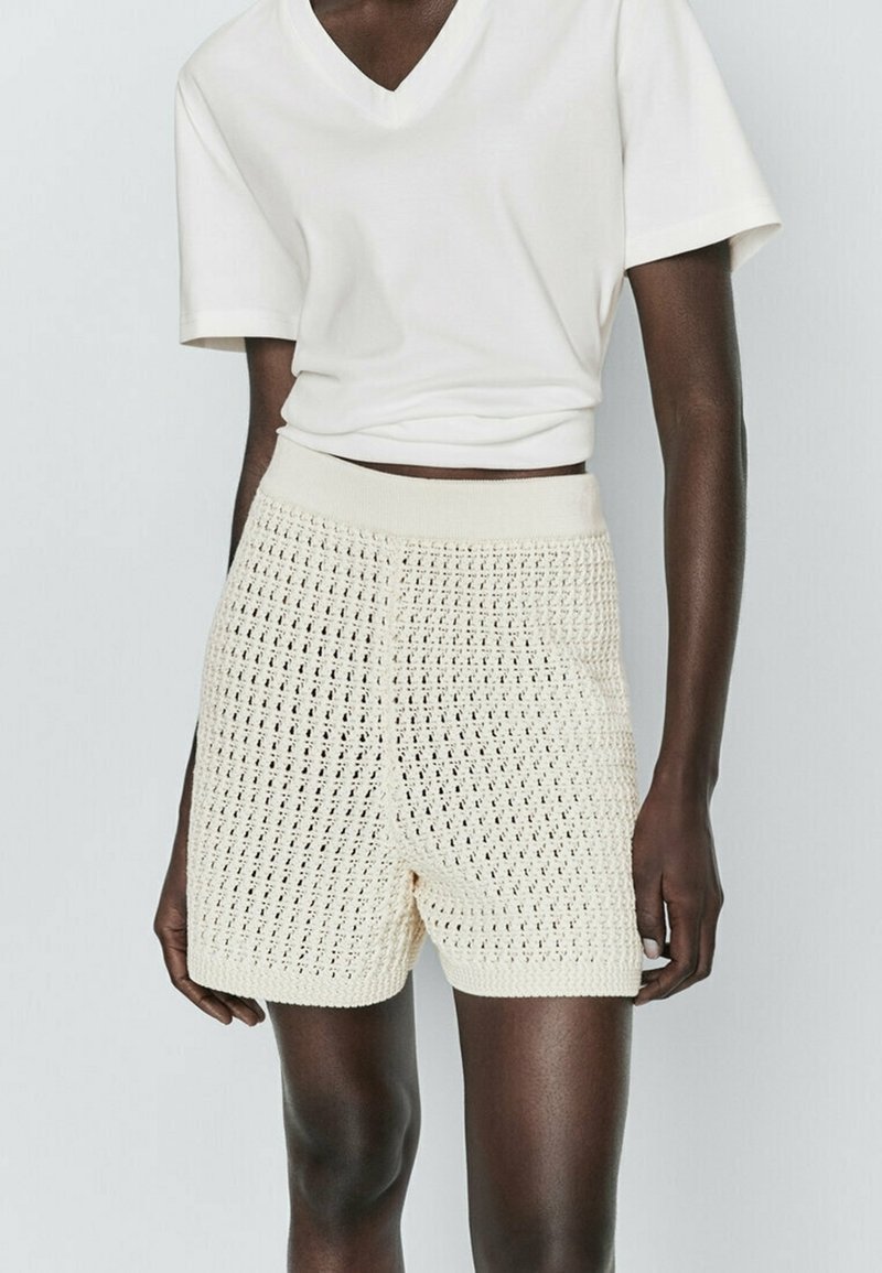 Cream-colored knitted shorts with a textured, open-weave pattern and elastic waistband, paired with a white cropped V-neck t-shirt.