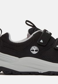 Chaussure de sport noire avec tige en mesh, sangles Velcro et accents gris. Présente un logo blanc et une semelle en caoutchouc texturé pour une meilleure traction.