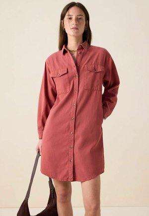 Button-up shirt dress in coral cotton with long sleeves, two chest pockets, and a curved hem. Model holding a dark handbag.