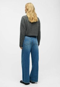 Mujer rubia vestida con un suéter gris oscuro y jeans de pierna ancha azul, de pie de espaldas contra un fondo claro y liso.