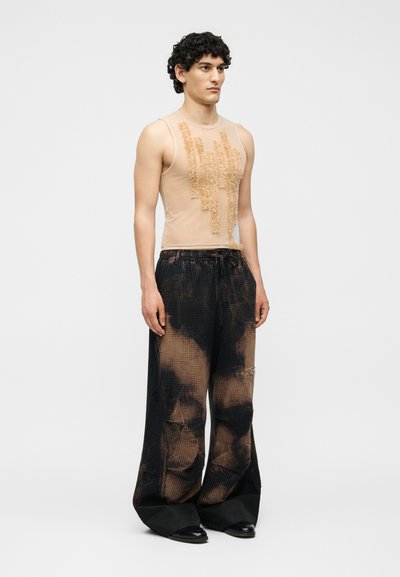 Νεαρός άνδρας στέκεται, φοράει μπεζ αμάνικη μπλούζα με υφασμάτινη λεπτομέρεια μπροστά και φαρδιά μαύρα παντελόνια με αποχρωματισμένα σχέδια.