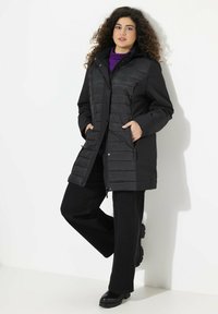 Cappotto imbottito nero con chiusura a zip e bottoni a pressione, e due tasche laterali. Abbinato a pantaloni neri e a un dolcevita viola sotto.