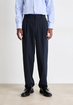 Pantaloni navy dal taglio rilassato, con una texture liscia, fronte plissettato e vita piatta, abbinati a scarpe nere lucide.