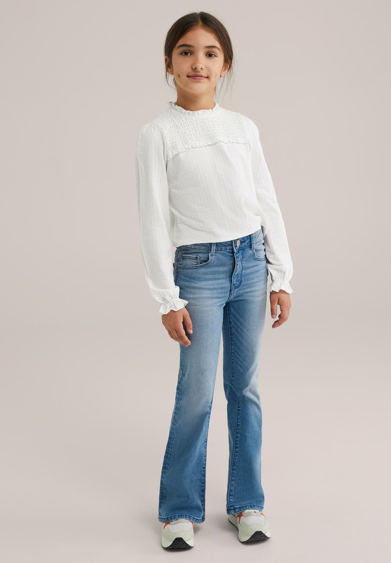 Top bianco a maniche lunghe con texture e dettaglio smock, abbinato a jeans chiari a zampa d'elefante e sneakers verdi con suola nera e accenti arancioni.