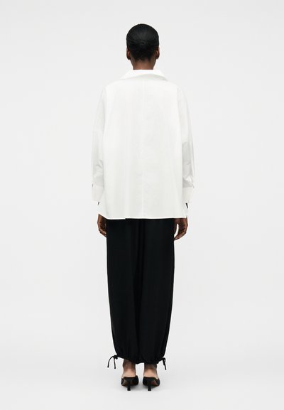 Žena s tmavou pokožkou a vlasy staženými do nízkého drdolu, oblékající volnou bílou košili s dlouhým rukávem a volné černé kalhoty, stojící zády k nám na bílém pozadí.