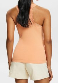 Camiseta de tirantes de melocotón con ribetes de encaje en las sisas, combinada con pantalones cortos en beige claro. El diseño ceñido y elegante resalta la espalda.