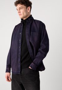 Cămașă din denim de culoare violet închis, cu nasturi pe față, un buzunar pe piept și un croi lejer, purtată deasupra unei bluze cu gât înalt negre.