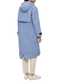 Persoon in een lichtblauwe met capuchon regenjas met rugventilatie, witte sneakers en beige cropped broeken, staande met de rug naar de kijker.