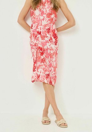 Donna che indossa un abito midi senza maniche con stampa floreale rosa e rossa, dettaglio arricciato, e sandali beige intrecciati, in piedi con le mani dietro la schiena.