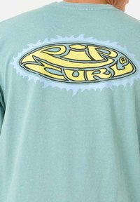 Hellblaue BaumwollT-Shirt mit einem großen gelben und schwarzen Rip Curl-Logo und einem Wellen-Design auf der Rückseite. Kurze Ärmel und Rundhalsausschnitt.