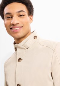 Beige jasje met een hoge kraag, voorzien van zichtbare bruine knopen. De stof lijkt glad en lichtgewicht, met een gestructureerde pasvorm.