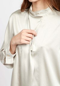 Blusa di seta grigia chiara con collo alto, dotata di un cordoncino con un pon-pon, maniche lunghe e polsini con bottoni.