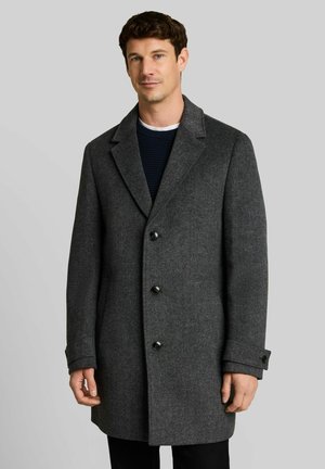 Uomo che indossa un soprabito grigio scuro con bottoni e un maglione nero, in piedi davanti a uno sfondo grigio chiaro uniforme.