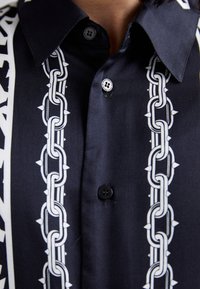 Μαύρο πουκάμισο με κοφτή κολάρο που διαθέτει λευκά σχέδια αλυσίδας με ακίδες, κουμπωμένο με δύο εμφανείς κουμπιά της Versace Jeans Couture.