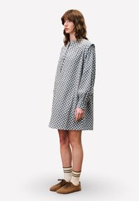 Karierte Bluse in Blau und Weiß, mit langen Ärmeln, lockerem Schnitt, knielang, mit einer teilweise durchgehenden Knopfleiste; kombiniert mit braunen Schuhen und gestreiften Socken.