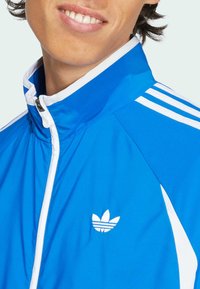 Blue athletic jacket with white accents, featuring a high collar and three stripes on the shoulders. Small white logo on the chest.