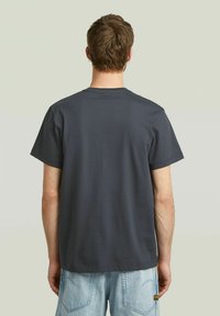 T-shirt di cotone grigio scuro con maniche corte, scollatura rotonda, texture liscia e senza grafiche o motivi visibili. Indossato con jeans azzurri chiari.