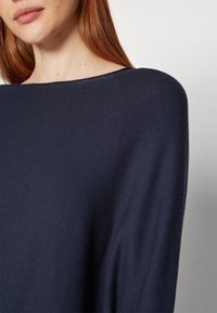 Pull en maille bleu marine à coupe ample, avec un large décolleté et une texture douce, présentant des coutures détaillées et une silhouette décontractée.