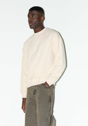 Mann mit dunkler Haut, der einen cremefarbenen Pullover und graue Jeans trägt, mit einer Hand in der Tasche stehend und nach links blickend vor einfachem Hintergrund.