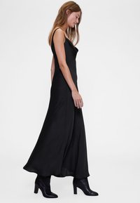 Melna maxidress bez piedurknēm ar dziļu izgriezumu, plūstošu audumu un A-veida siluetu, kombinēta ar melniem potīšu zābakiem ar papēdi.
