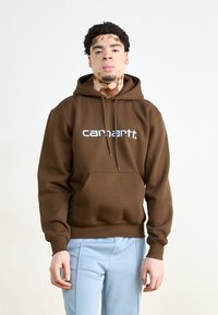Brūns kapuce krekls ar priekšējo kabatu un baltu "carhartt." logo. Gluda tekstūra un brīva piegriezuma, kopā ar gaiši ziliem biksēm.