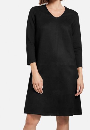 Femme portant une robe noire à manches longues, longueur genou, avec un col en V et une jupe légèrement évasée, debout devant un fond blanc.